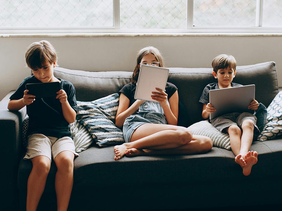 Three children sat on a sofa using computers