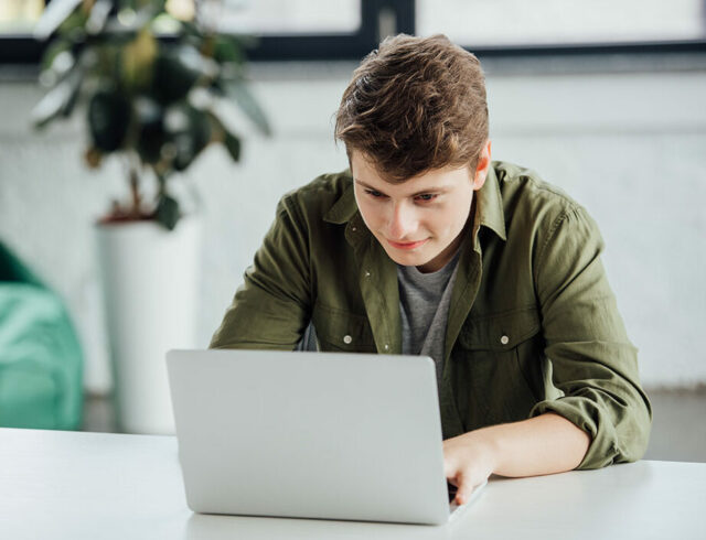 smiling teen boy sitting at table and using laptop