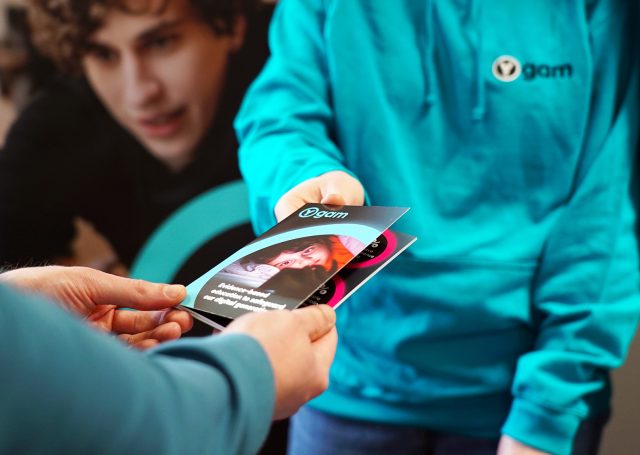 A Ygam staff member at an event is handing a Ygam brochure to a passer by.